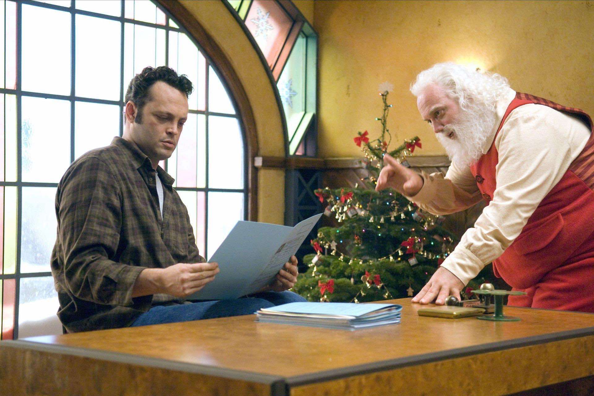 Image of Fred Claus sitting at a desk reviewing whether children are naughty or nice with his brother Santa standing next to him.